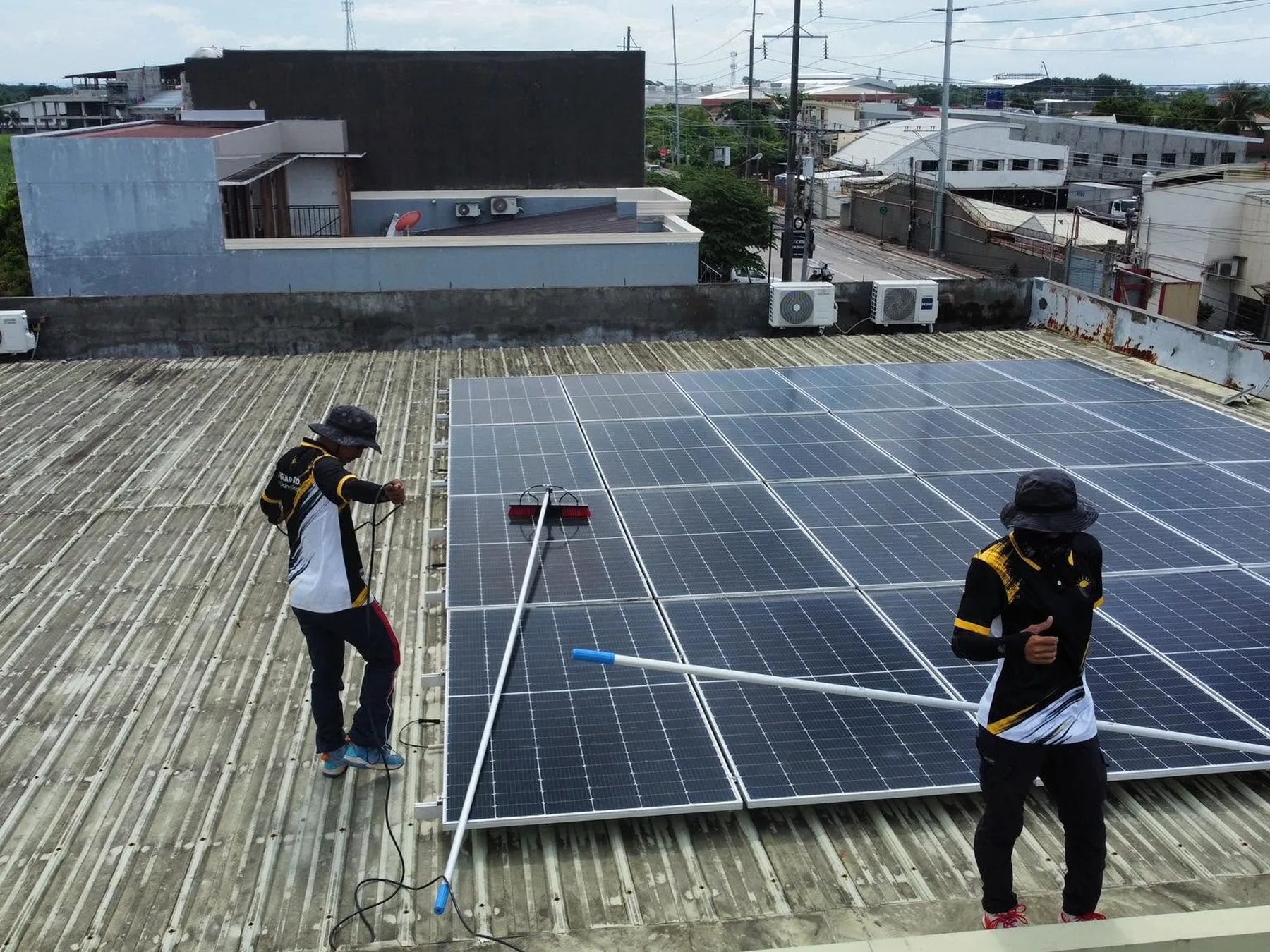 Two technicians cleaning a large rooftop solar array with water-fed brush poles.